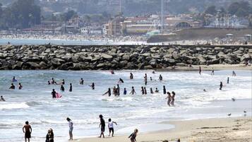 A crowded beach with people swimming and walking along the shore, a rocky breakwater in the background, and buildings on the far side.
