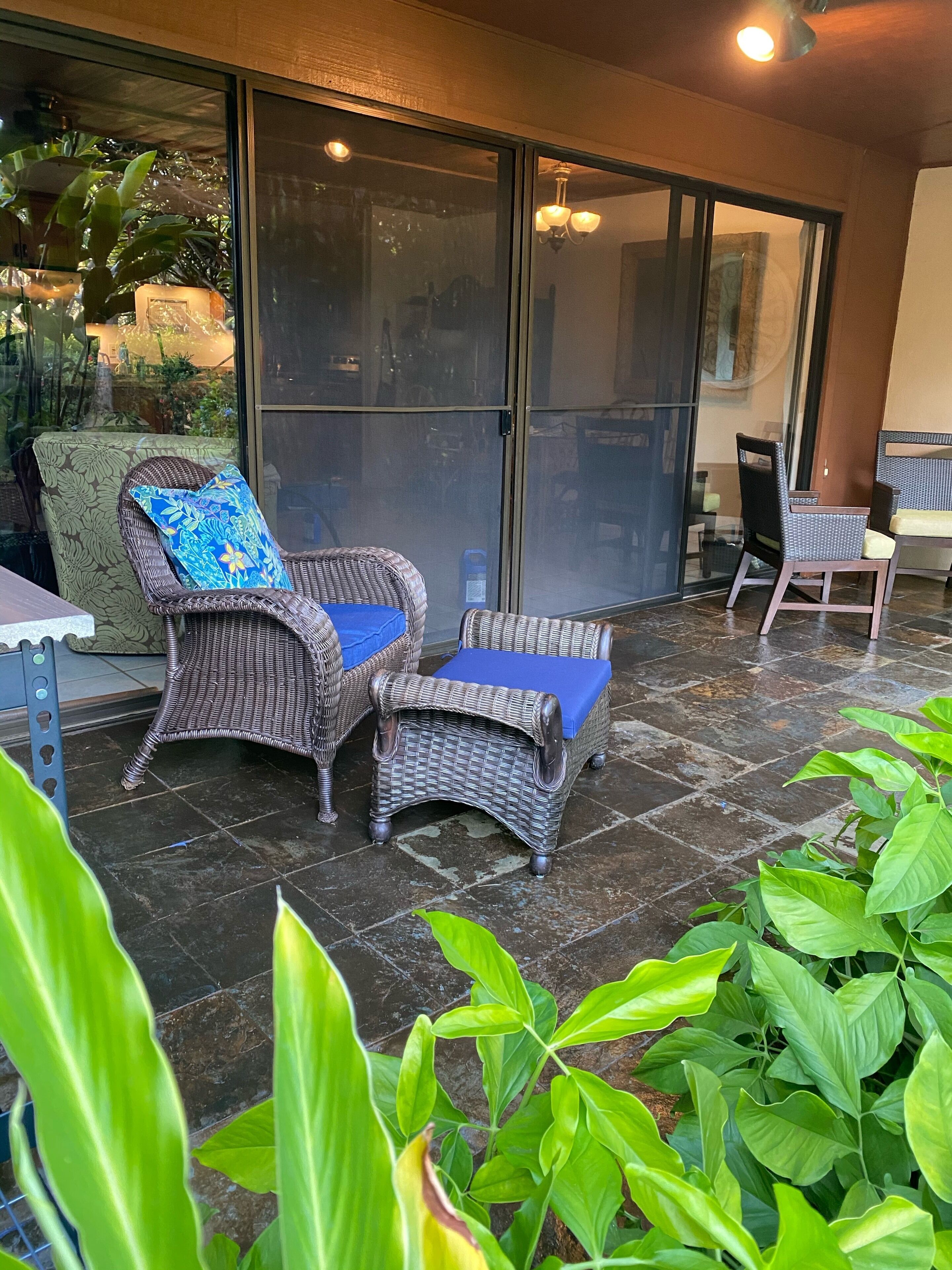 Un patio techado con mobiliario de mimbre, incluyendo dos sillones y una mesa, y una puerta de vidrio que da a un espacio al aire libre.