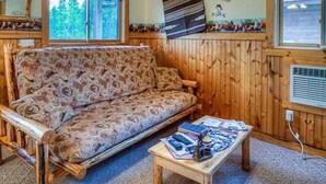A cozy cabin interior with a sofa, a small table, and a window overlooking greenery. - #3 - COWBOY CABIN – BEAUTIFUL PRIVATE SETTING, 6 MILES TO GLACIER PARK (Columbia Falls)
