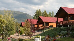 A row of wooden cabins with red roofs, a balcony, and a garden with flowers and shrubs. - #3 - COWBOY CABIN – BEAUTIFUL PRIVATE SETTING, 6 MILES TO GLACIER PARK (Columbia Falls)