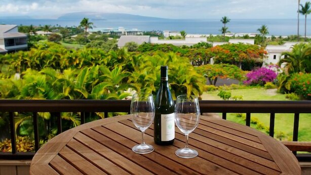 A wooden table with a bottle of wine and two glasses on a balcony overlooking a lush garden and the ocean. - #708 Wailea Ekolu - Spectacular Ocean View, Serene Setting (Wailea-Makena)