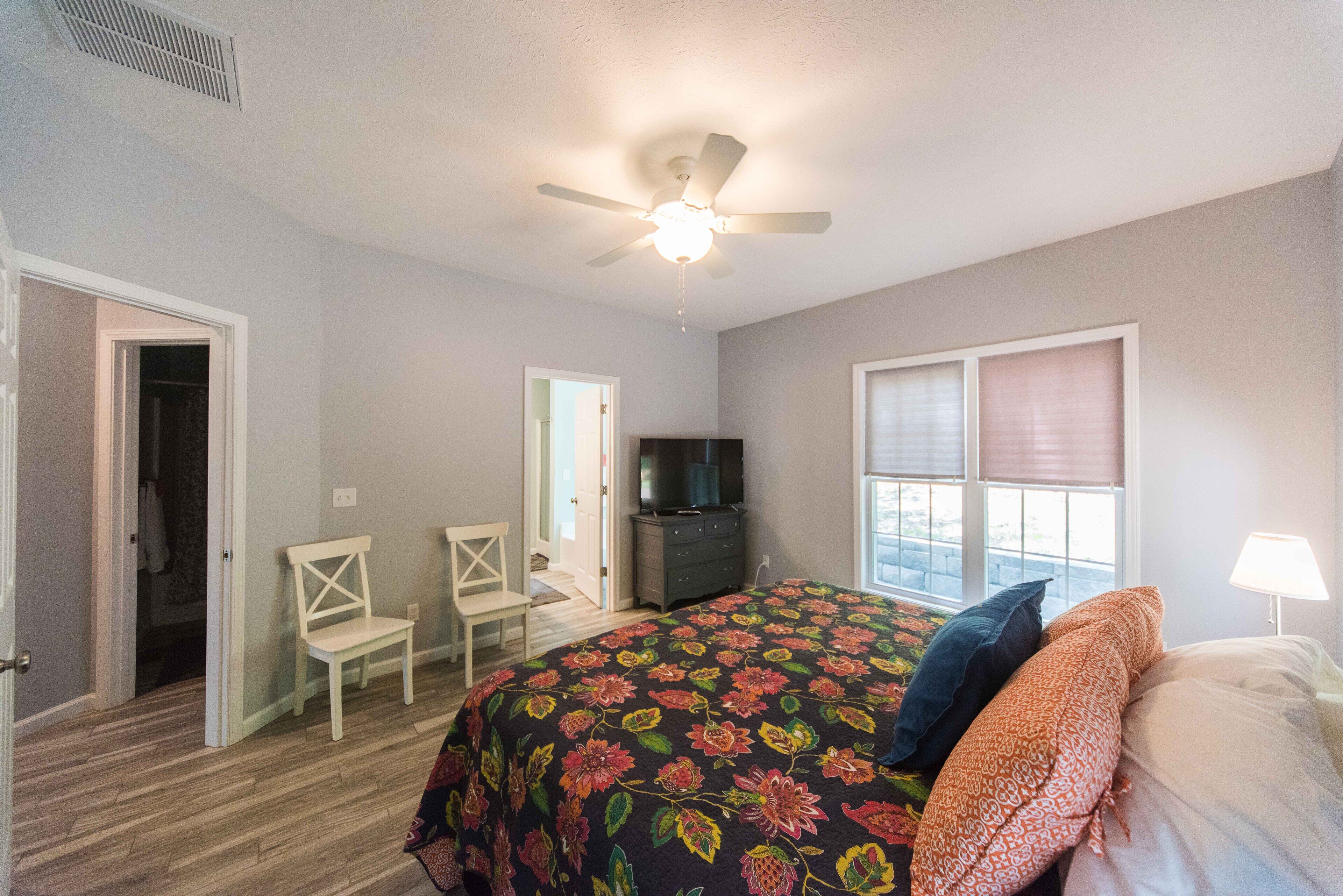 A bedroom with a bed, a ceiling fan, a TV, a chair, and a window with blinds.