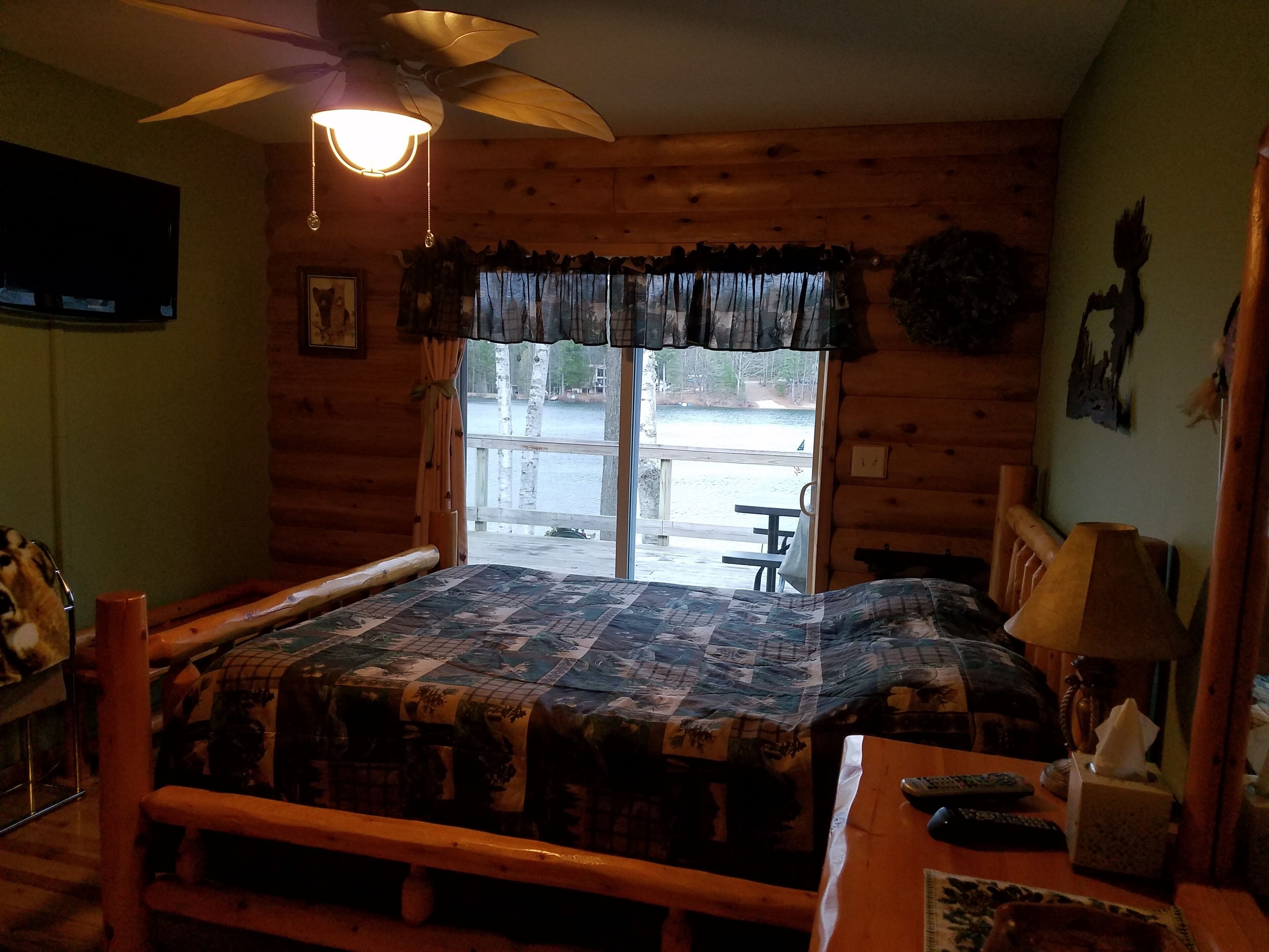 Een slaapkamer in een blokhut met een bed, een bureau, een stoel, een plafondventilator en een raam met uitzicht op een steiger en water.