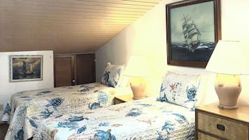 Une chambre avec un plafond en pente, deux lits avec la même literie à motifs floraux, une table de chevet avec une lampe, une commode en bois et un tableau encadré représentant un navire accroché au mur.