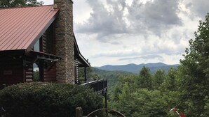 A rustic cabin with a stone chimney, a wooden deck, and a view of a mountainous landscape.