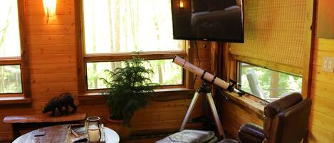 A cozy cabin interior with a wooden interior, a mounted telescope, a comfortable leather chair, a small table with a candle, and a television on the wall.
