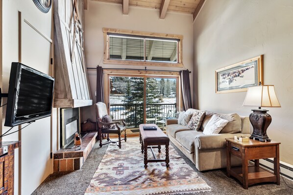 A cozy living room with a fireplace, a television, a sofa, a coffee table, and a window with a view of trees.