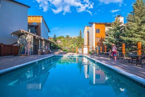 A swimming pool with a wooden deck, surrounded by buildings and trees.