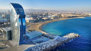 Un edificio moderno con fachada de vidrio curva cerca de una zona costera, con mar azul cristalino y un rompeolas rocoso.