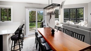 A modern kitchen with a wooden table and bar stools, large windows, and a balcony with a view of trees. - Coldstream Townhome 15 | Spacious 5BR Vail Townhome (Vail)