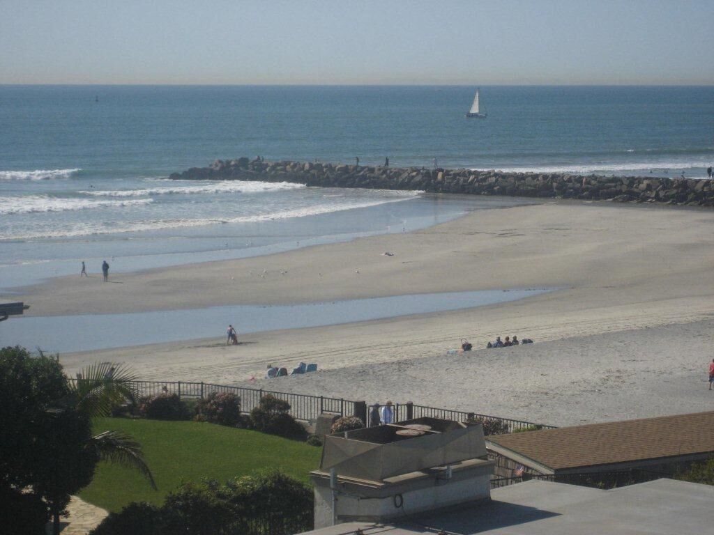 A beach with a rocky breakwater, a sailboat in the distance, and people walking along the shore.