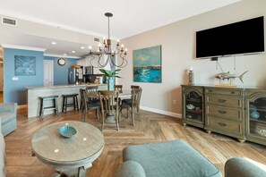 A modern living room with a dining area, wooden flooring, a chandelier, a flat-screen TV, and a painting of a seascape.