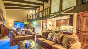 A spacious living room with a large brown leather sofa, a coffee table with books and a remote control, and a staircase leading to an upper level.