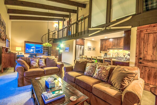 A spacious living room with a large brown leather sofa, a coffee table with books and a remote control, and a staircase leading to an upper level.