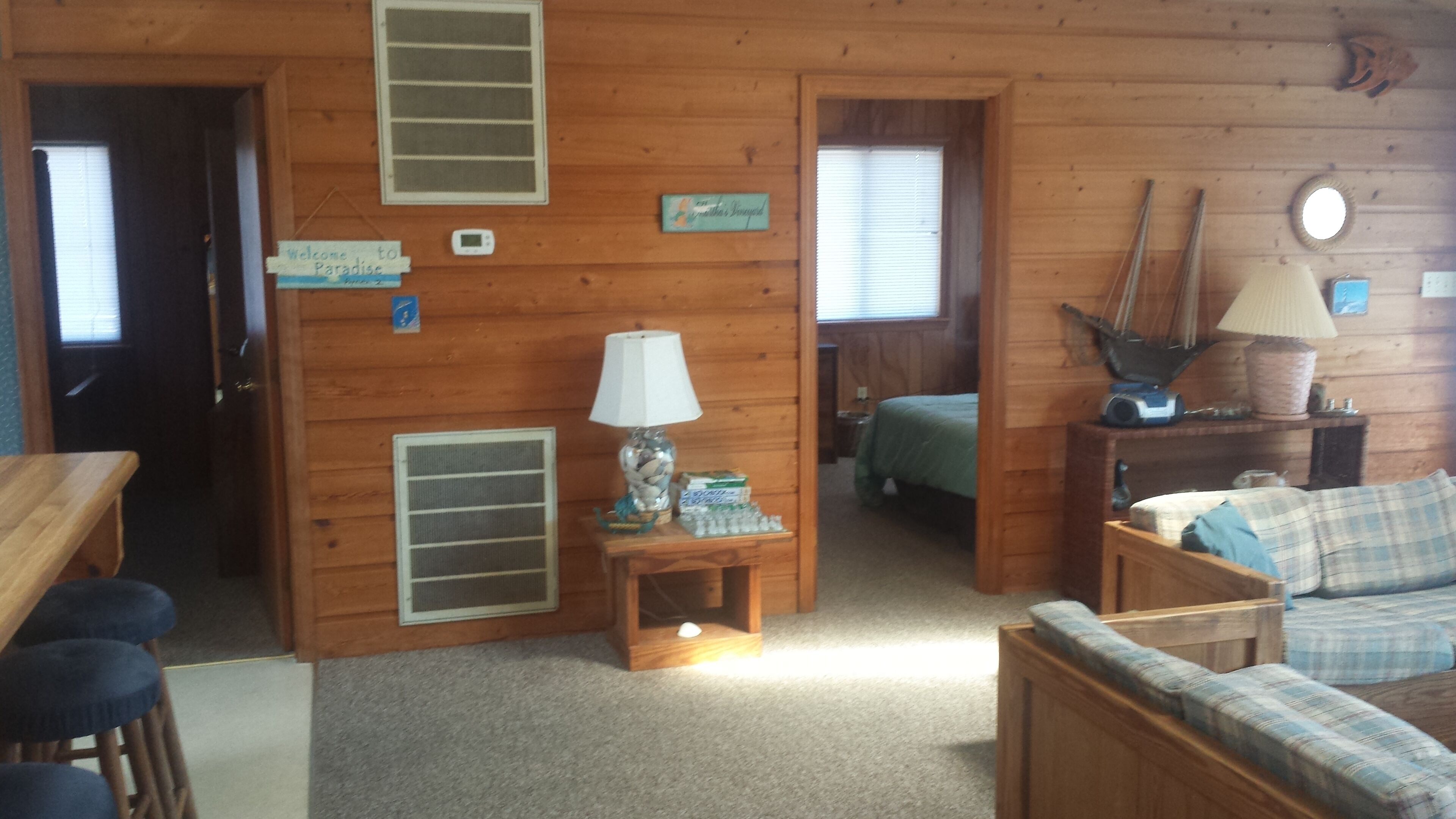 A wooden cabin interior with a dining area, a living room with a sofa and a lamp, a bed, and a small table with a lamp and decorative items.
