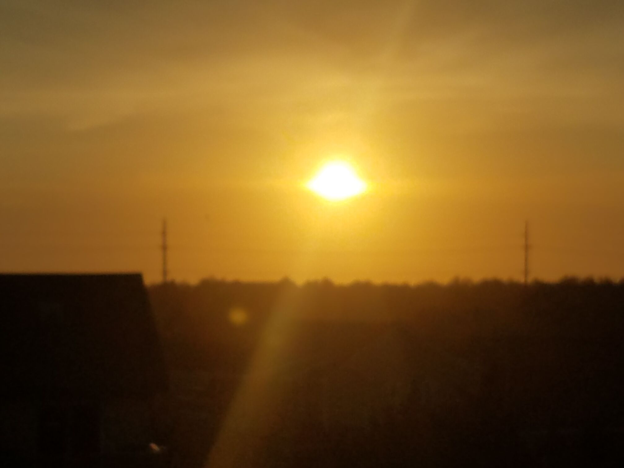 A sunset with the sun low on the horizon, casting a warm glow over a silhouette of buildings and a power line structure.