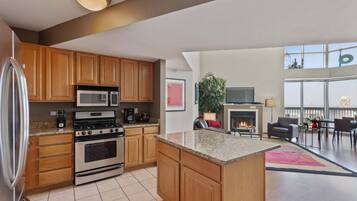 A modern kitchen with wooden cabinets, a stainless steel refrigerator, a microwave, and an oven. There is a dining area with a fireplace, a TV, and a balcony with a view.