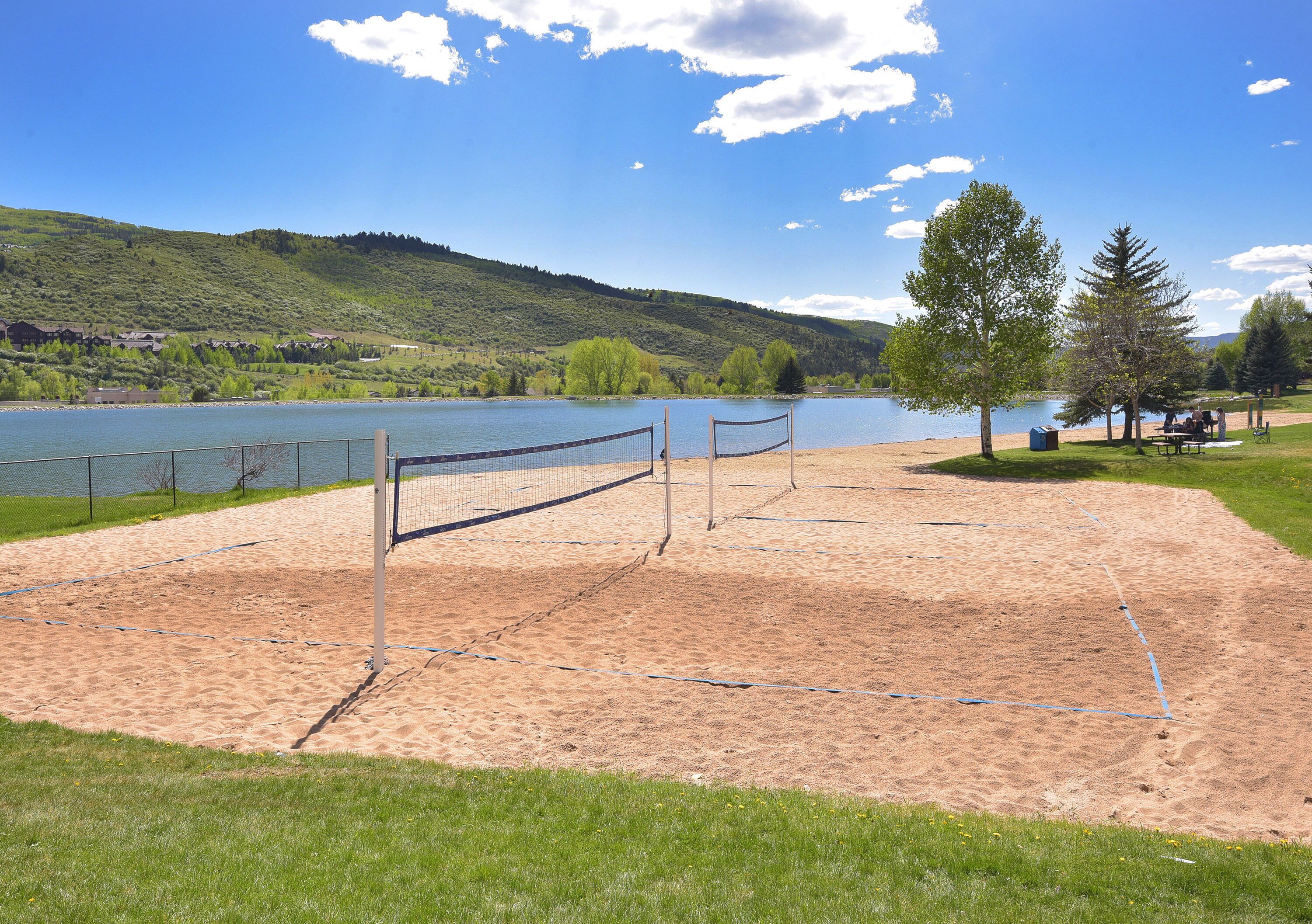 A sandy beach volleyball court with a net, a lake, and trees in the background.