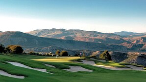 A golf course with sand bunkers, a putting green, and a backdrop of mountains.