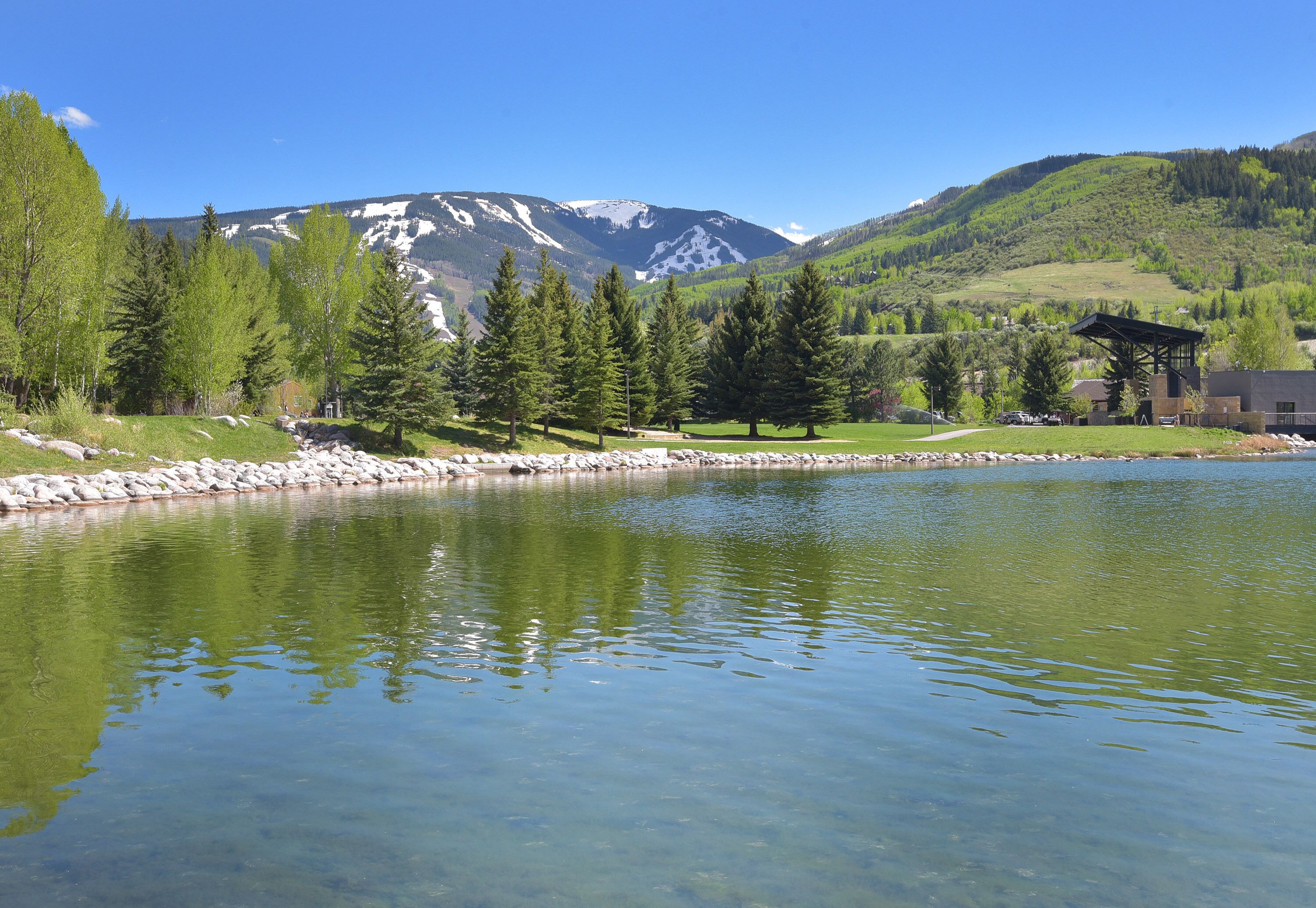 A clear lake with a rocky shoreline, surrounded by evergreen trees and a mountain backdrop.