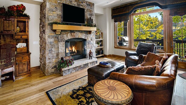 A living room with a stone fireplace, leather furniture, and a large window with a view of trees. - Beaver Creek Landing 206 | 3BR Ski-In/Ski-Out, Summer AC (Beaver Creek)