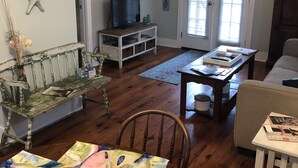 A cozy living room with a wooden floor, a television, a bench, a table with a colorful tablecloth, and decorative starfish on the wall. - Quaint Cottage, Walk to Beach and Old Town Bay St. Louis (Bay St Louis)