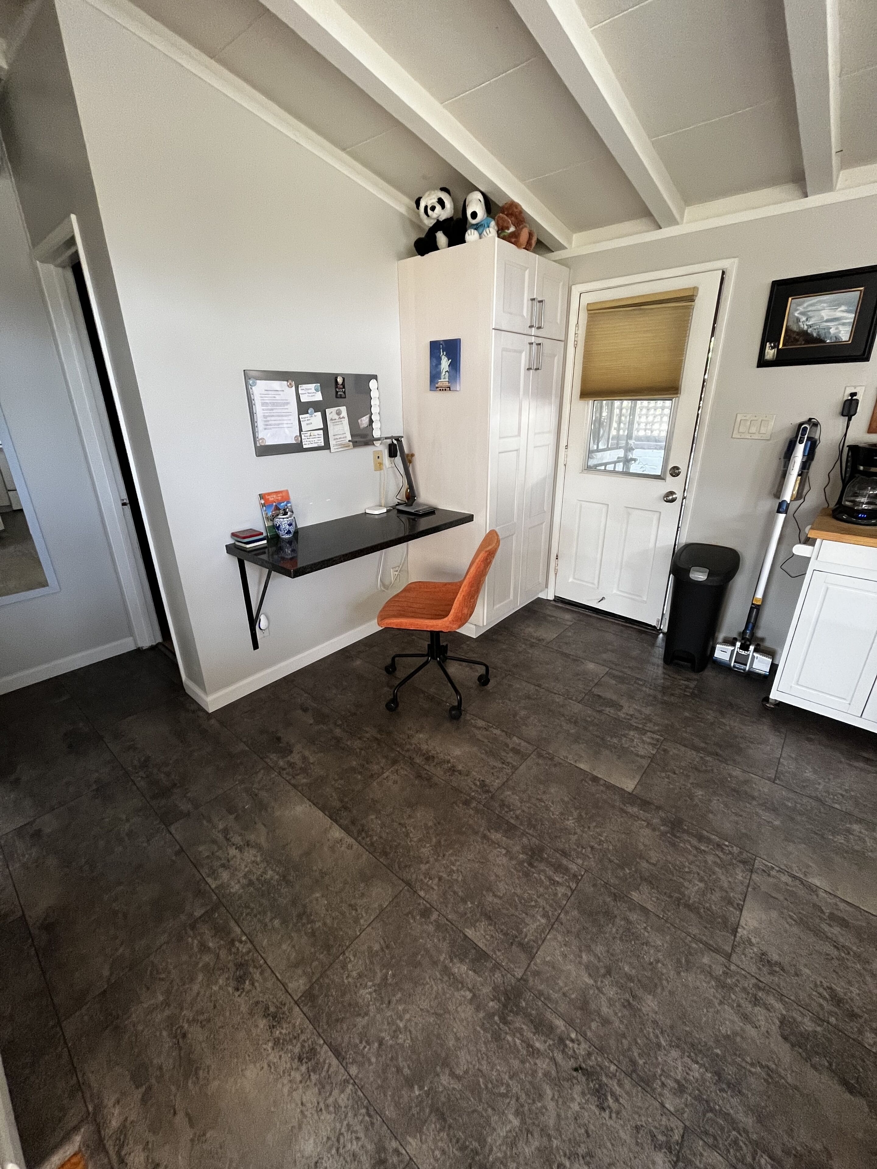 A home office with a desk, chair, and storage cabinets. There is a window with blinds, a trash can, and a vacuum cleaner.