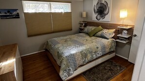 Un dormitorio con cama, ventana con persianas, mesita de noche con lámpara y un cuadro en la pared.