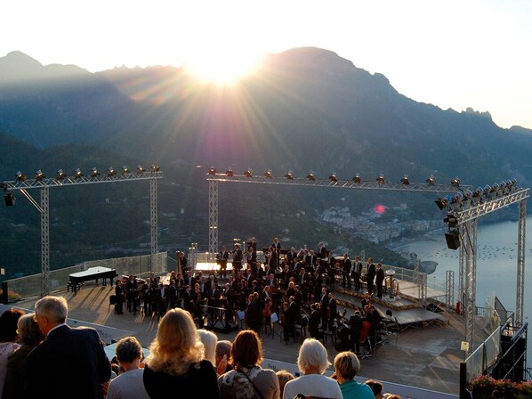 An outdoor concert stage with a crowd of people, surrounded by mountains and a body of water.