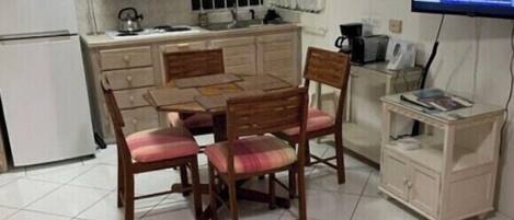 Un espacio habitable pequeño y funcional con kitchenette, una zona de comedor con mesa y sillas de madera, un televisor y un mueble pequeño.