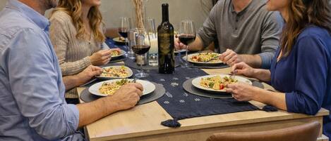 A group of people dining at a table with a bottle of wine, plates of food, and a neon sign in the background.