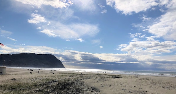 A beach with people walking along the shore, a lighthouse in the background, and a clear sky with clouds.