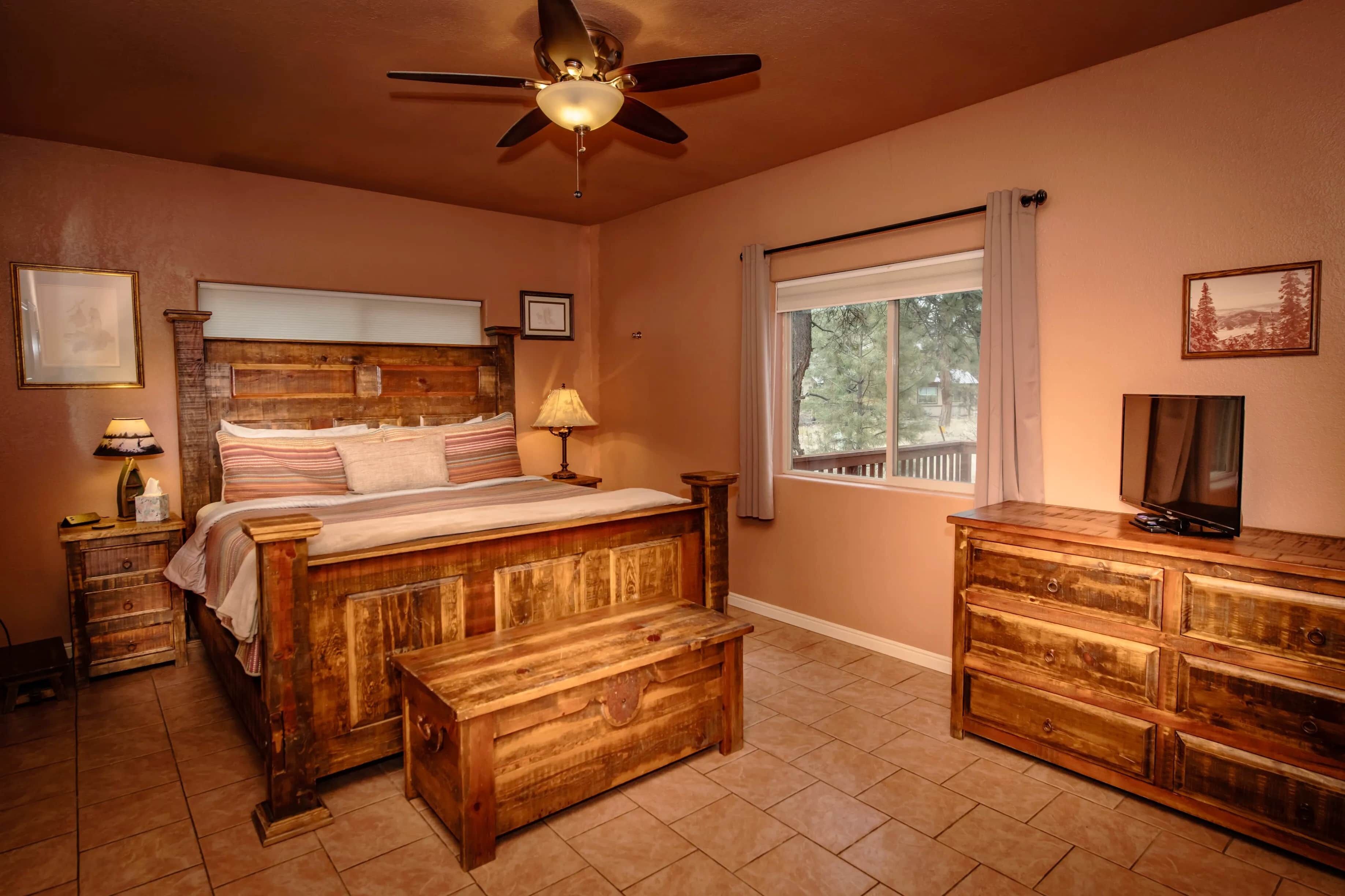Un dormitorio con una cama de madera, una mesita de noche, un aparador, un ventilador de techo y una ventana con vistas a árboles.
