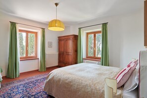 A bedroom with a large bed, two windows with curtains, a radiator, and a wooden wardrobe.