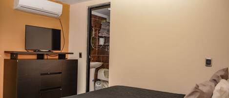 A hotel room with a bed, a TV on a stand, an air conditioner, and a bathroom visible through an open door.