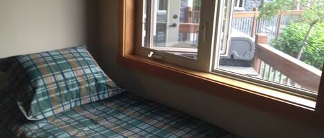 A small, cozy room with a single bed featuring a plaid bedspread. A window provides a view of a balcony with outdoor furniture and greenery.