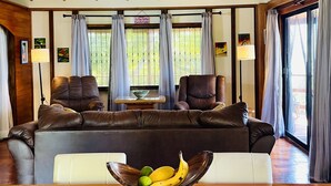 A living room with a wooden ceiling, leather sofas, a dining table with a fruit bowl, and a view through large windows.