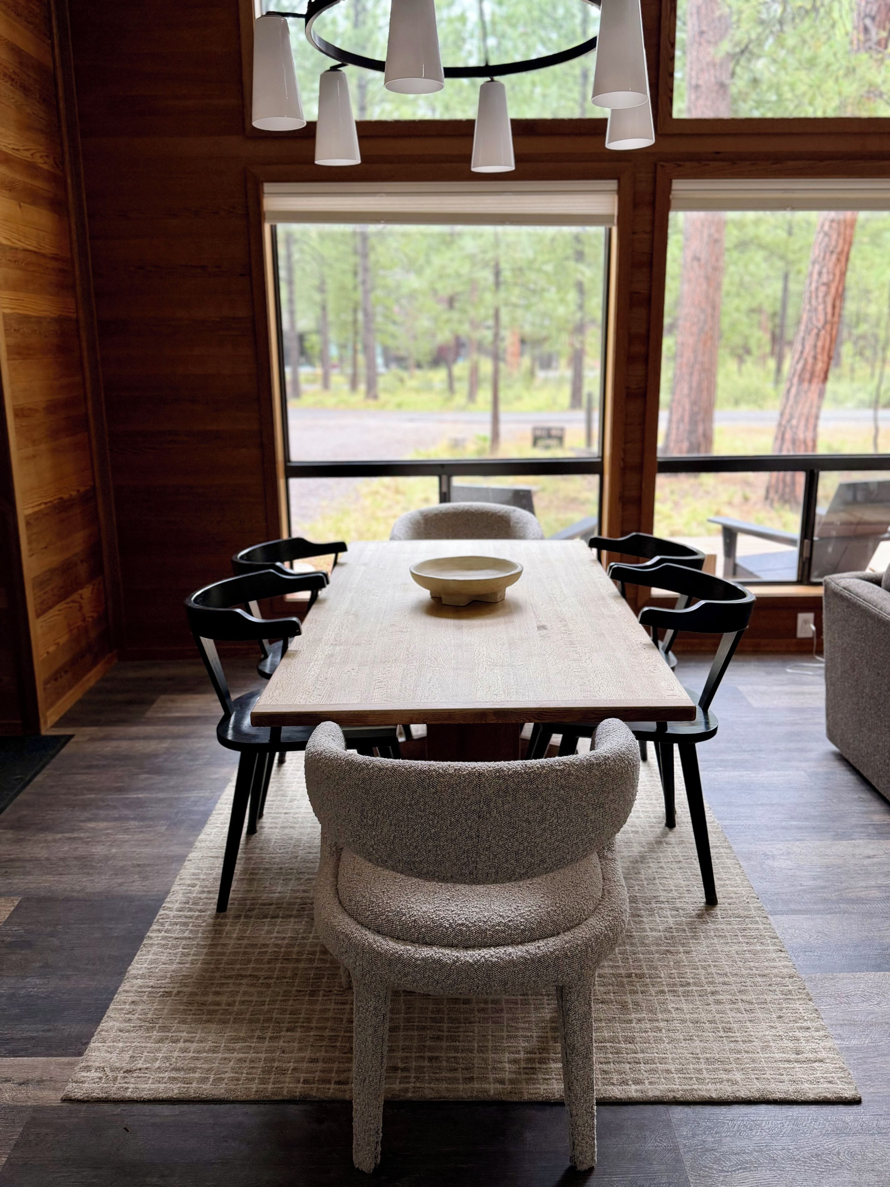 A dining area with a wooden table, six chairs, a bowl, and a view of the outdoors through large windows.