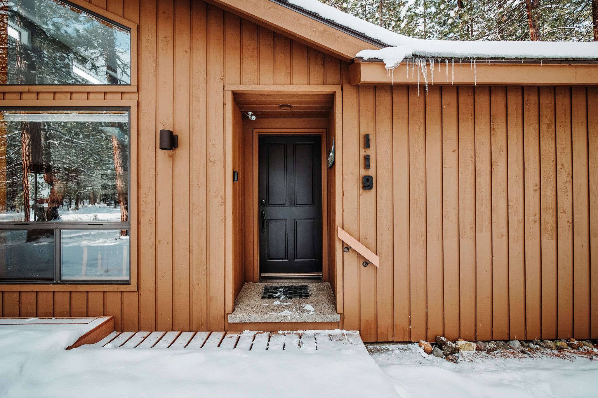 A wooden cabin with a black door, a small porch, and a window overlooking a snowy landscape.