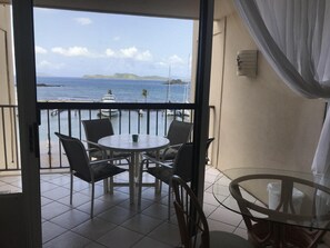 A balcony with a view of the sea, a table with chairs, and a glass table on the right.