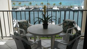 Een balkon met een ronde tafel, vier stoelen, een bloempot en uitzicht op een jachthaven met boten en palmbomen.