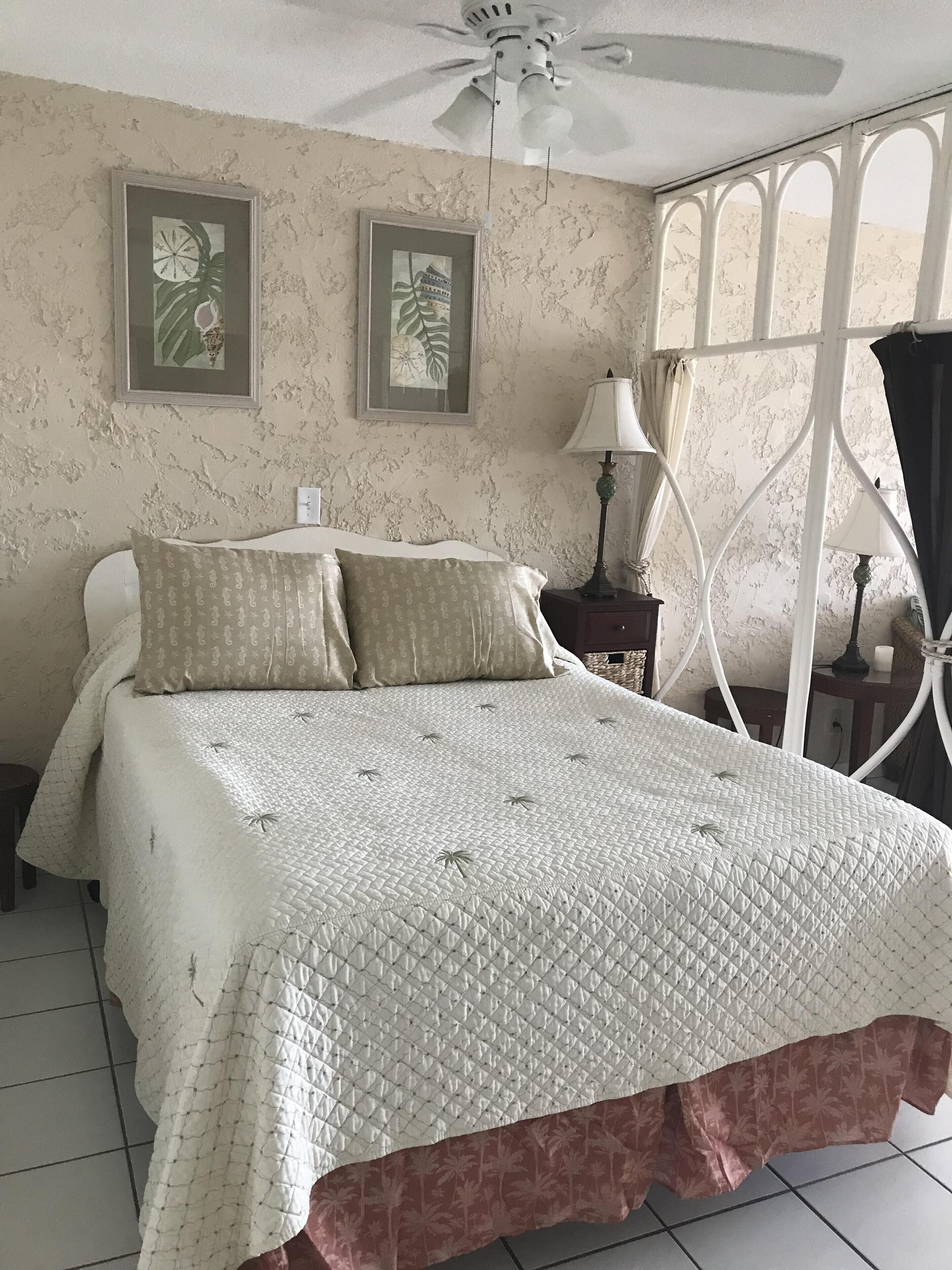 A bedroom with a bed, a ceiling fan, two framed pictures on the wall, a nightstand with a lamp, and a window with curtains.