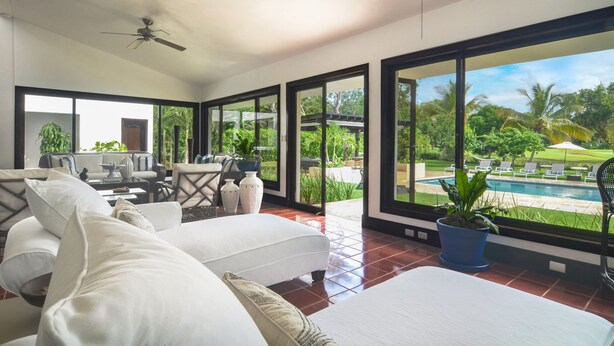 Una habitación amplia con un gran ventanal, ventilador de techo, sofá y vista a una piscina.