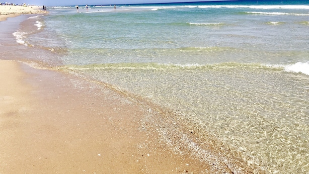 Ein Strand mit klarem, flachem Wasser, sandigem Ufer und einigen Personen in der Ferne.