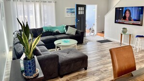 Ein Wohnzimmer mit einem dunkelgrauen Sofa, einem runden Couchtisch und einem braunen Ledersessel. An der Wand ist ein großer Fernseher angebracht, der einen Nachrichtensender zeigt.
