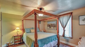 A four-poster bed with a wooden frame, a bedside table with a lamp, a framed picture on the wall, and a window with curtains.