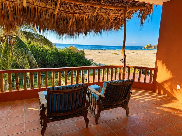 Eine Terrasse mit zwei Holzstühlen und einem Tisch, mit Blick auf einen Strand mit Palmen und dem Meer.