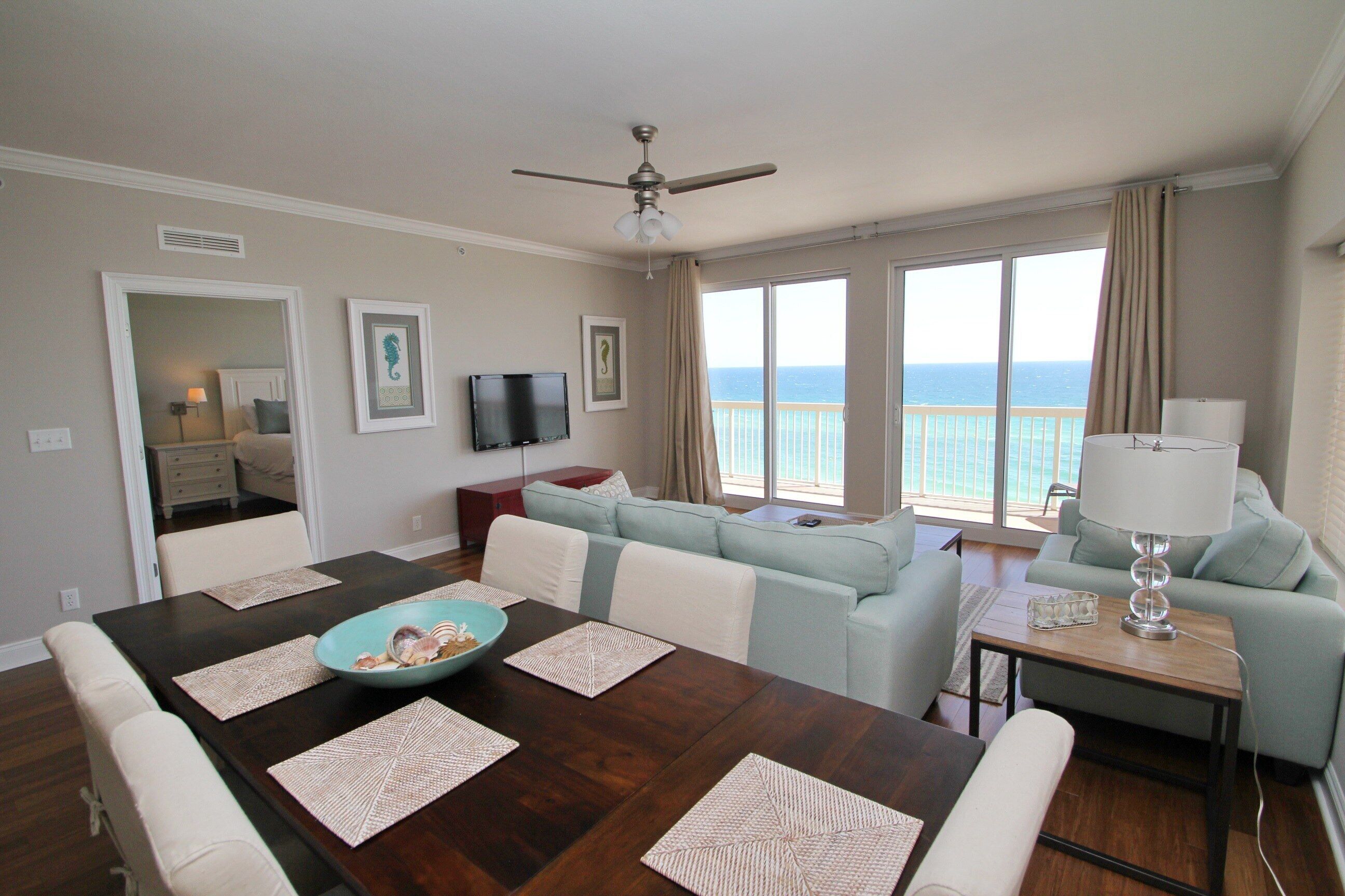 A dining area with a dark wooden table, white chairs, and a blue bowl. There is a light blue sofa and a small table with a lamp. A ceiling fan is visible, and the room has large windows with a view of the ocean.