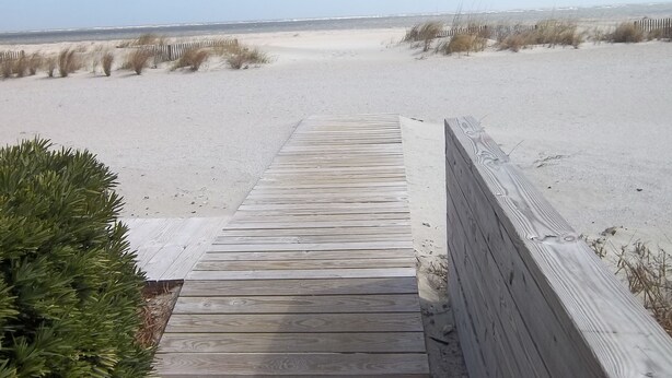 En træpromenade fører til en sandstrand med klitter og sparsom vegetation.