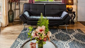 Een woonkamer met een zwarte leren bank, een groot raam met gordijnen, een vloerlamp en een vaas met bloemen op een kamerkleed.